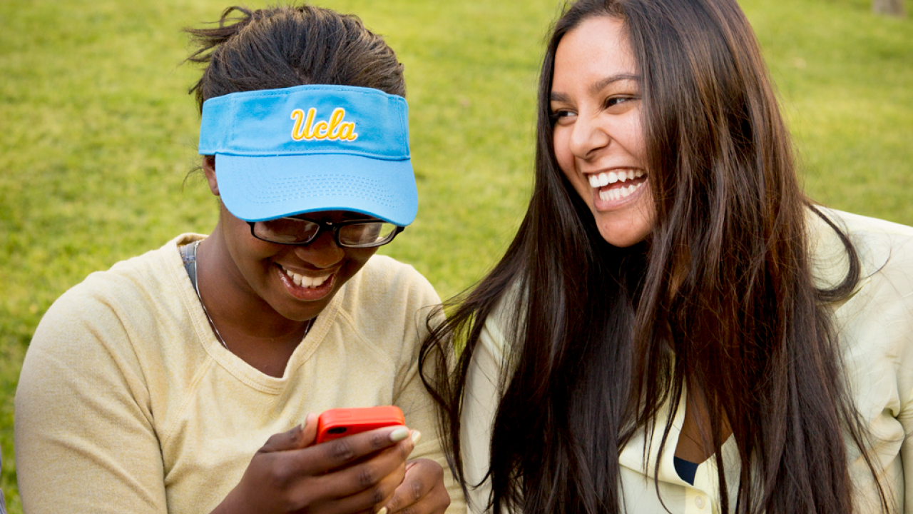Two students laugh while sitting on the edge of a grassed area. The student on the left is wearing a UCLA visor and is looking at her phone. 