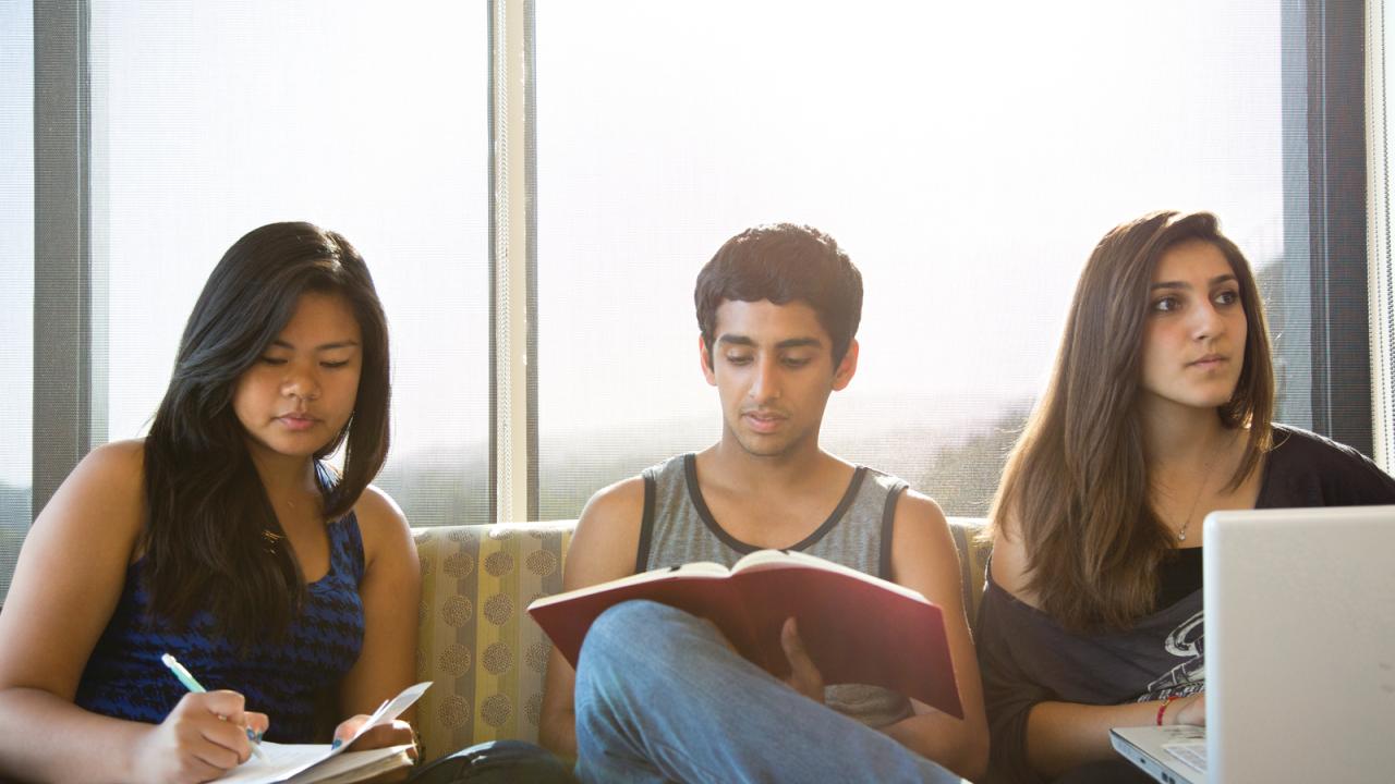 Three students sit in front of a window, taking notes on their laptops and in notebooks.