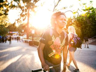 A student wearing a 'UCLA Black Bruin' t-shirt pushes their scooter up Bruin Walk.