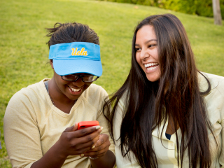 Two students laugh while sitting on the edge of a grassed area. The student on the left is wearing a UCLA visor and is looking at her phone. 