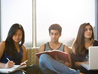 Three students sit in front of a window, taking notes on their laptops and in notebooks.